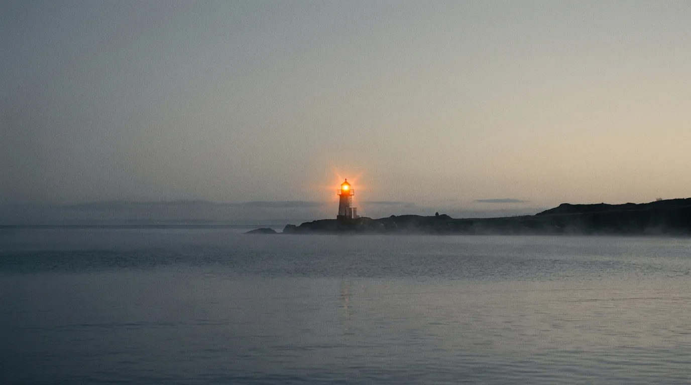A coastal light switching on as the horizon lightens over still water.
