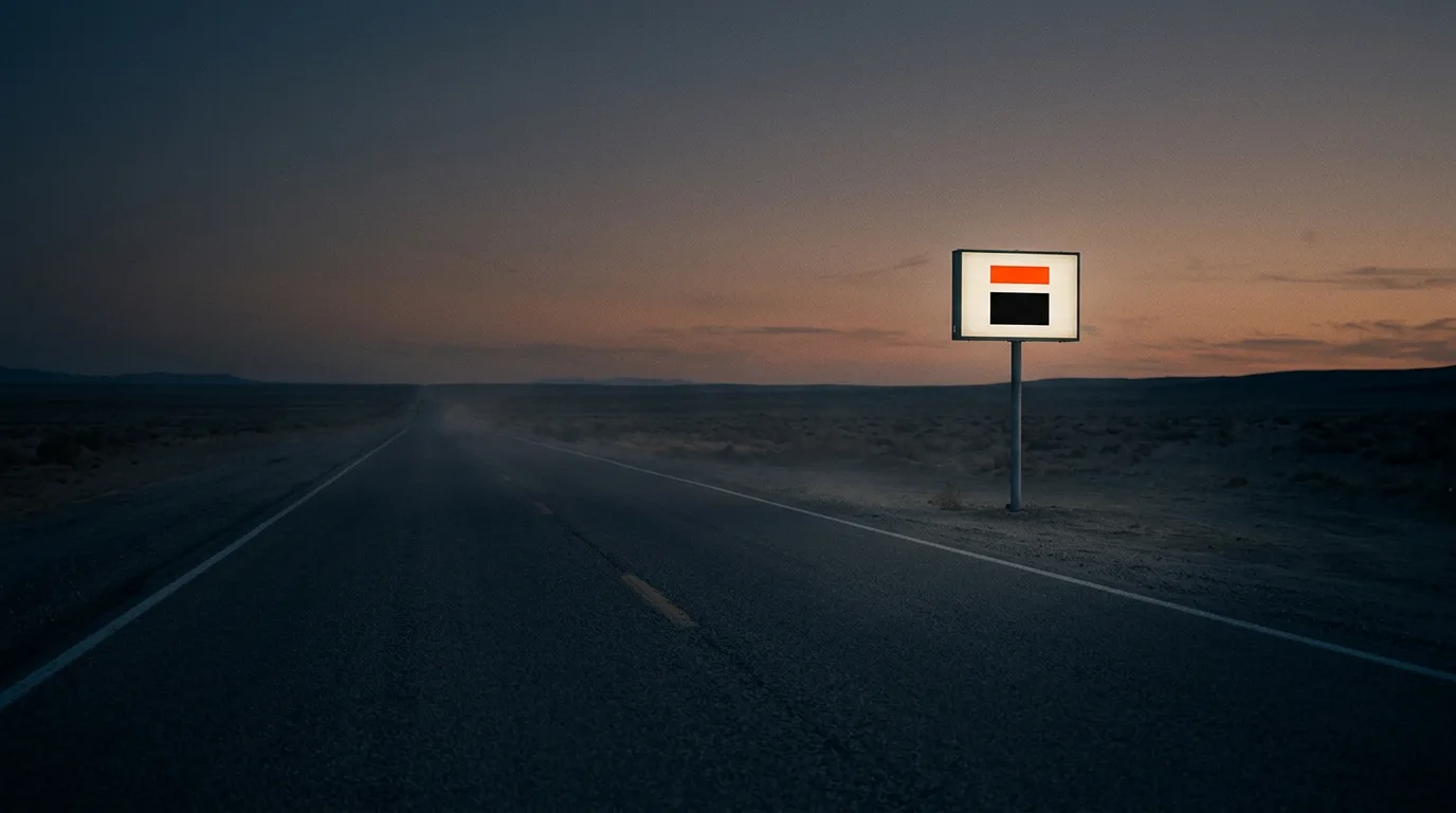 A long empty two-lane road stretching toward the horizon at dusk, one roadside sign lit from within, a small mark on its face.
