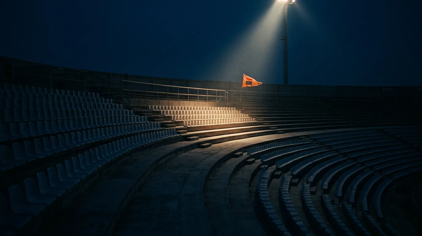 Empty stadium terraces fade into shadow except for one section lit by a single floodlight.